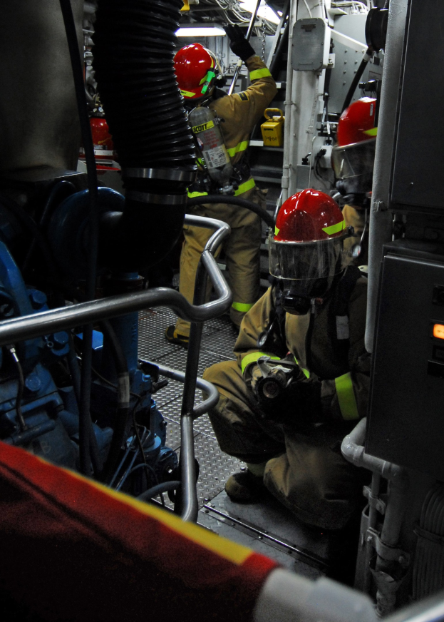 Feuerwehrleute in Helmen und Schutzausrüstung arbeiten an einem Feuerlöschwagen mit Rohren und einer Leuchte im Hintergrund.