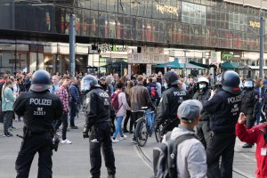 Eine Gruppe von Polizisten steht vor einer Menge auf der Straße, einige tragen Helme und haben Taschen oder halten Fahrräder, im Hintergrund sind Gebäude, Laternenmäste und Schirme zu sehen.