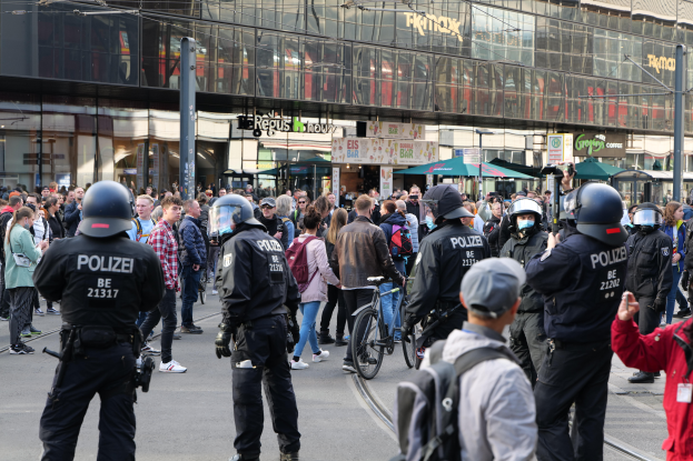 Eine Gruppe von Polizisten steht vor einer Menge auf der Straße, einige tragen Helme und haben Taschen oder halten Fahrräder, im Hintergrund sind Gebäude, Laternenmäste und Schirme zu sehen.
