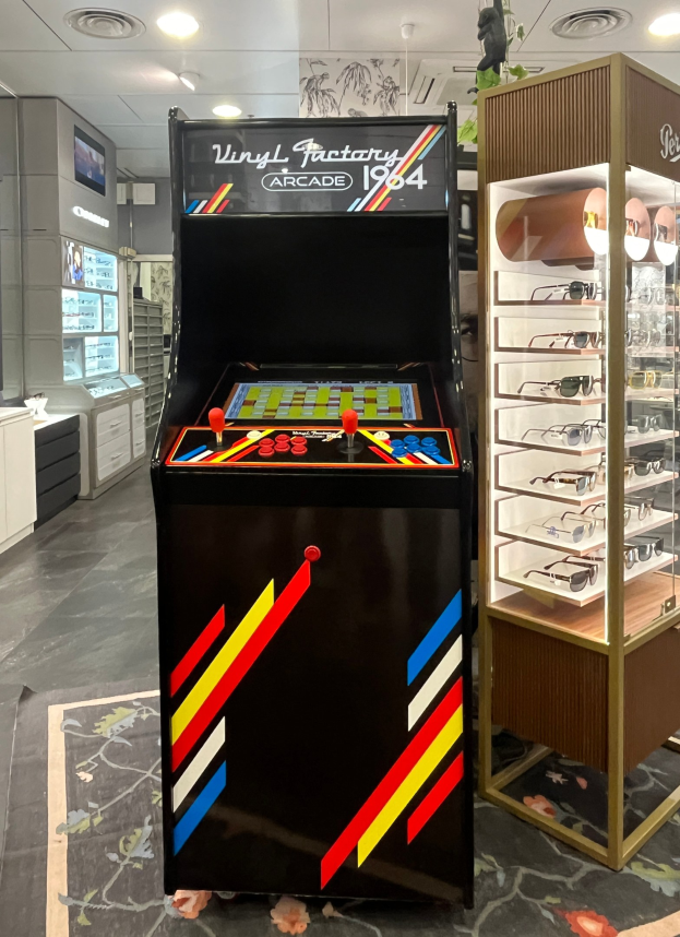 Ein Geschäft mit einem zentralen Retro-Arcade-Automaten, einer Glasvitrine auf der rechten Seite und einer retro-stylischen Beleuchtung mit Schränken, Fernseher und anderen Gegenständen im Hintergrund.