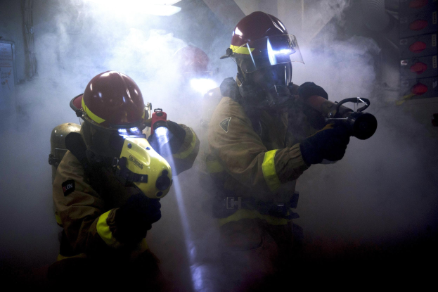 Zwei Feuerwehrleute in Schutzausrüstung sprühen Wasser auf eine rauchgefüllte Wand, mit einer Tafel mit Text links und verschiedenen Gegenständen rechts.