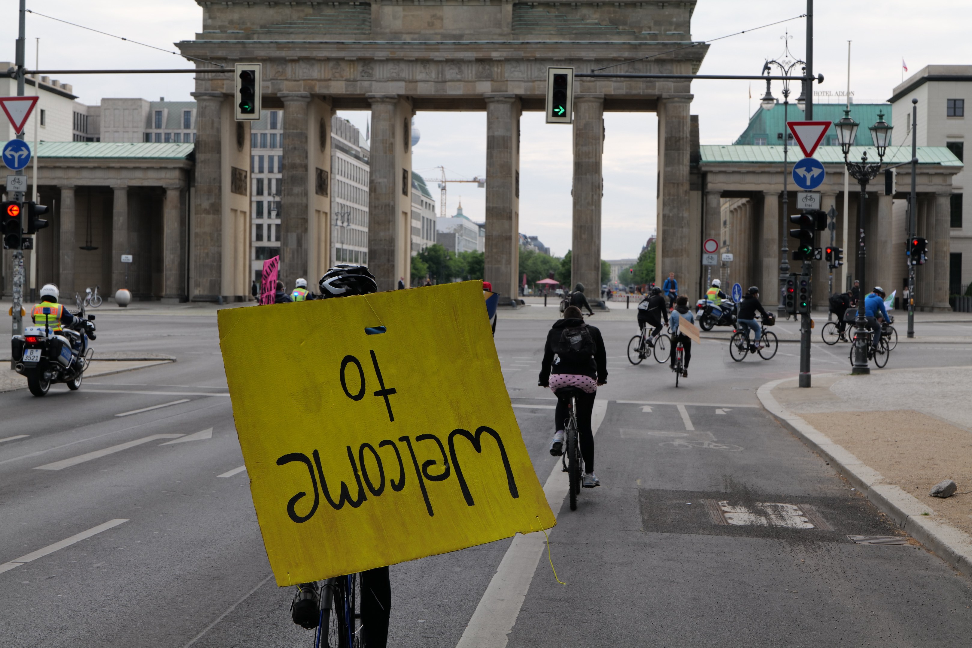 Eine Gruppe von Radfahrern fährt an der Brandenburgertor in Berlin vorbei, einer hält ein gelbes Schild, mit Laternenmasten, Verkehrszeichen, Gebäuden und Bäumen im Hintergrund unter einem klaren Himmel.