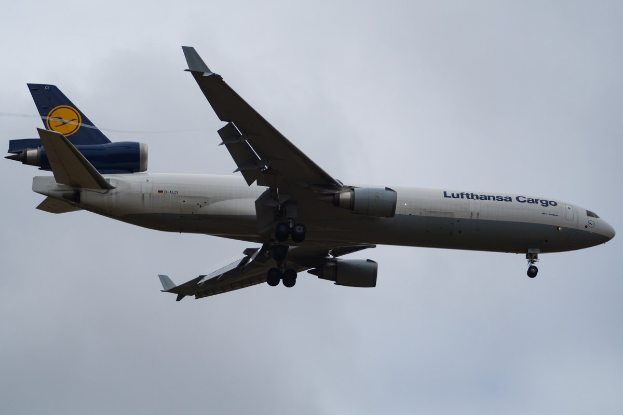 Lufthansa Cargo Boeing 777-300ER im Flug mit sichtbarem Text auf dem Rumpf, ausgefahrenen Tragflächen und weißen Wolken im Hintergrund.