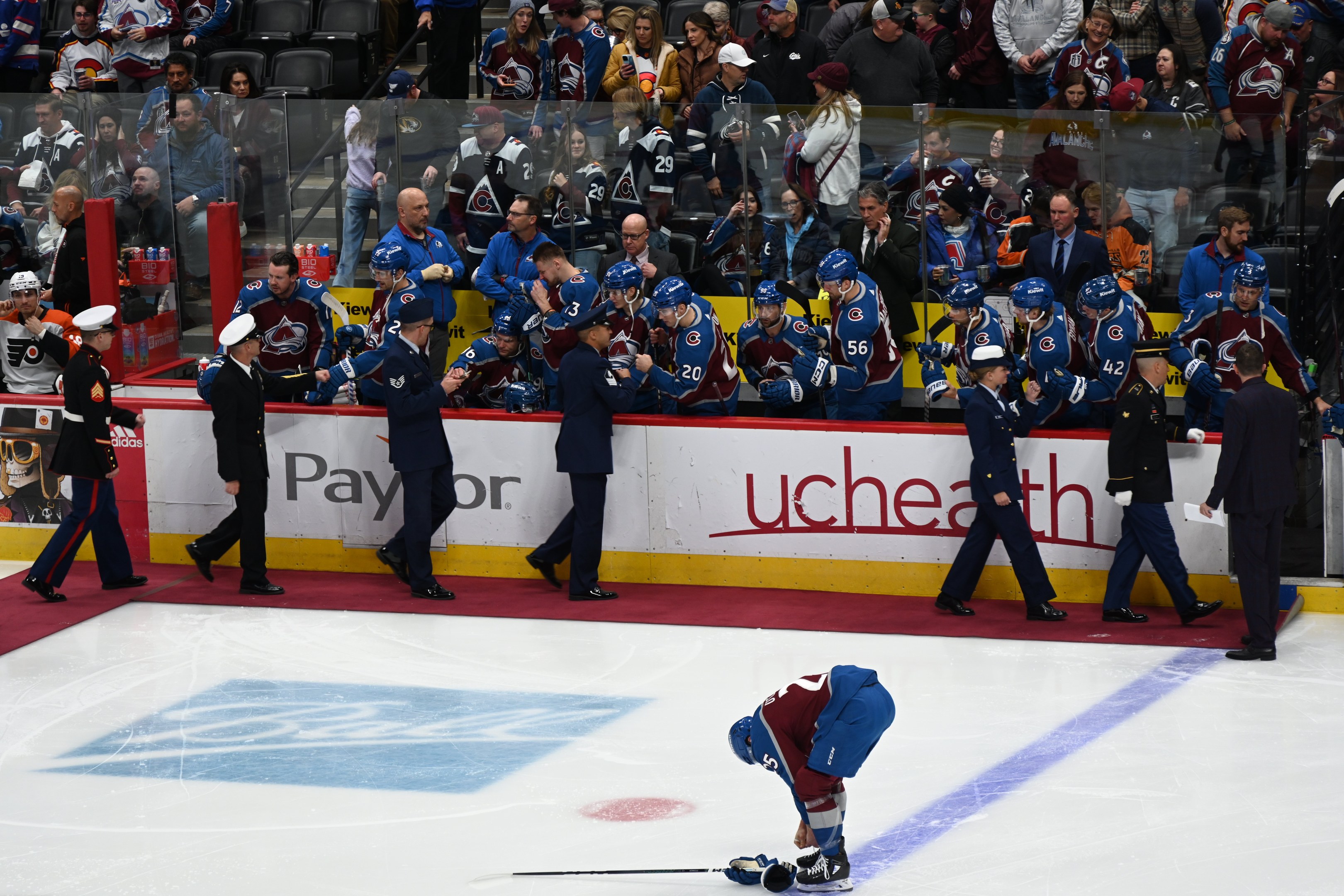Eishockeyspieler auf dem Eis umgeben von Menschen mit Mützen und Eishockeyschlägern, mit Zuschauern im Hintergrund und Werbetafeln "Colorado Avalanche vs San Jose Sharks - 3/24/15 NHL Pick, Odds, and Prediction".