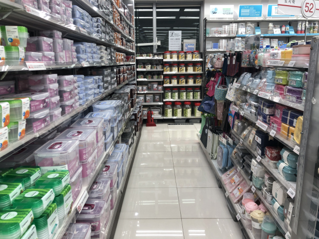 Ein unordentlicher chinesischer Supermarktgang mit Regalen, die mit Kartons, Flaschen, Taschen und anderen Produkten gefüllt sind, beleuchtet von Deckenlampen, mit Informationsschildern, einem Feuerlöscher und chinesischen Schriftzeichen.