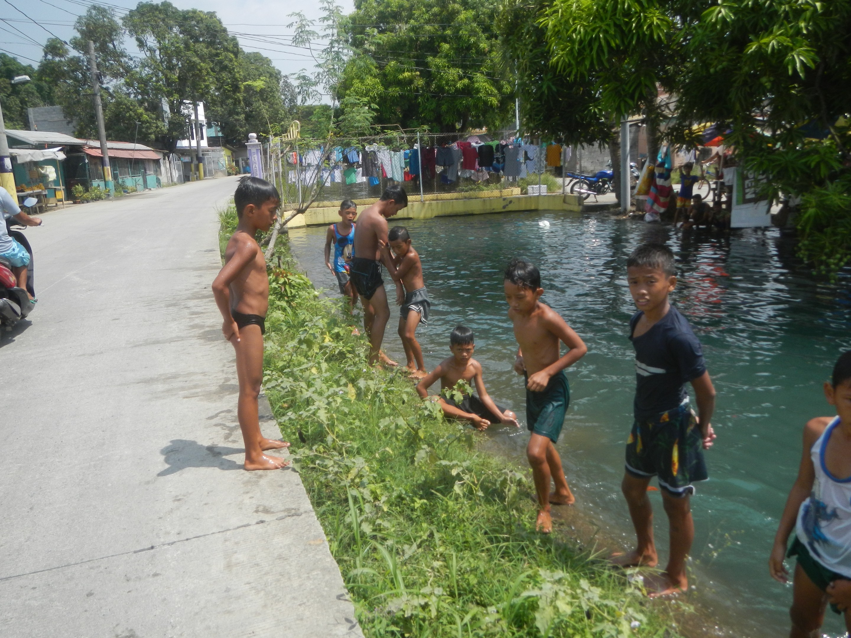 Kinder spielen in flachem Wasser neben einer Straße, einige stehen, während andere auf dem Gras sitzen, mit einer Person auf einem Motorrad links und Häusern, Bäumen und Strommasten im Hintergrund unter einem klaren blauen Himmel.