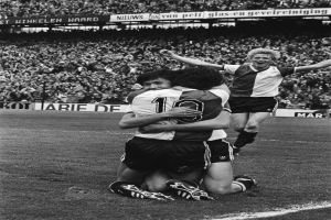 Drei Fussballspieler umarmen sich auf dem Boden und feiern ein Tor, während eine jubelnde Menge und schwenkende Fahnen im schwarz-weissen Hintergrund zu sehen sind.