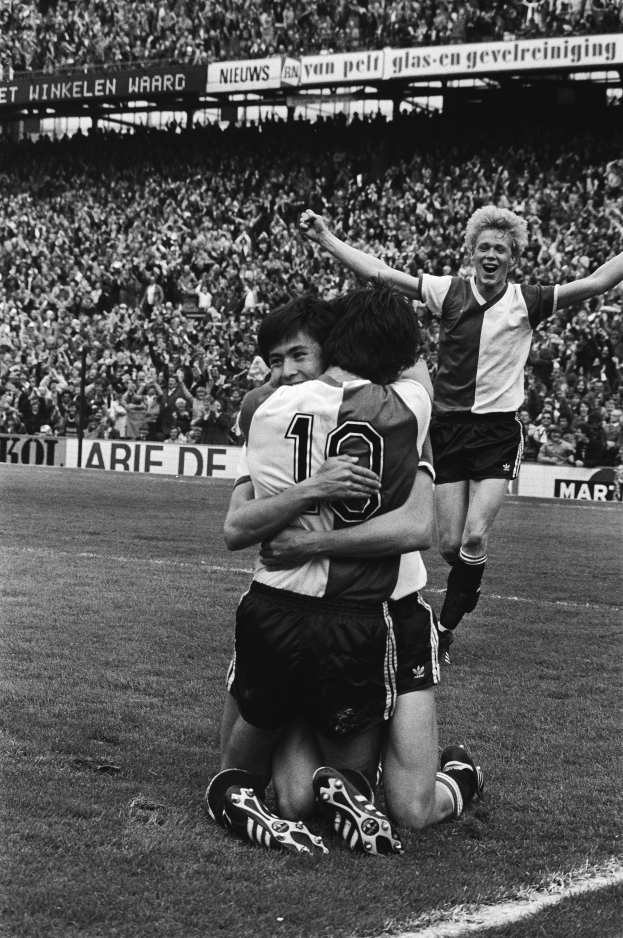 Drei Fussballspieler umarmen sich auf dem Boden und feiern ein Tor, während eine jubelnde Menge und schwenkende Fahnen im schwarz-weissen Hintergrund zu sehen sind.