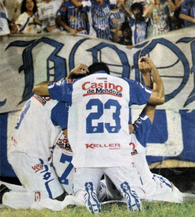 Eine Gruppe von Fußballspielern feiert ein Tor auf dem Feld mit jubelnden Zuschauern im Hintergrund und einem teilweise sichtbaren Banner mit Text.