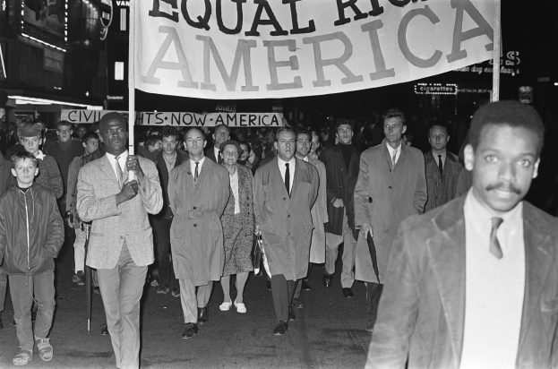 Schwarz-weiß-Foto einer Gruppe von Menschen, die eine Straße entlang marschieren und ein Banner mit der Aufschrift "Gleiche Rechte Amerika" tragen, mit Gebäuden und Straßenlaternen am Straßenrand.
