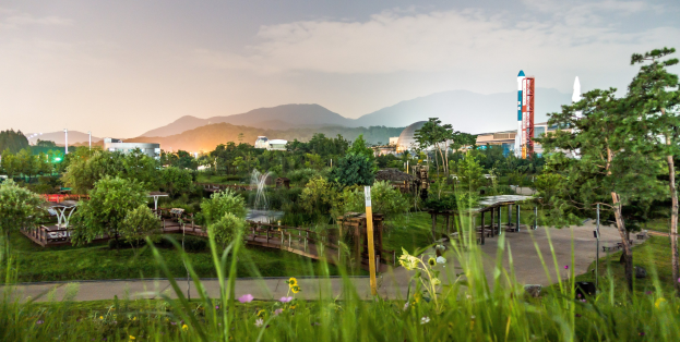 Ein Park mit grünem Gras, bunten Blumen, hohen Bäumen, einer Brücke, einem Brunnen, Laternenmasten und Gebäuden, vor Bergen und einem blauen Himmel.