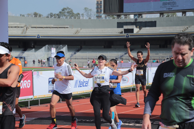 Gruppe von Menschen, die auf einer Stadionbahn laufen, mit Bannern, einer Anzeigetafel, einem Zaun, einem Gebäude, Bäumen und einem bewölkten Himmel; einige Läufer tragen Turnschuhe und Hüte.