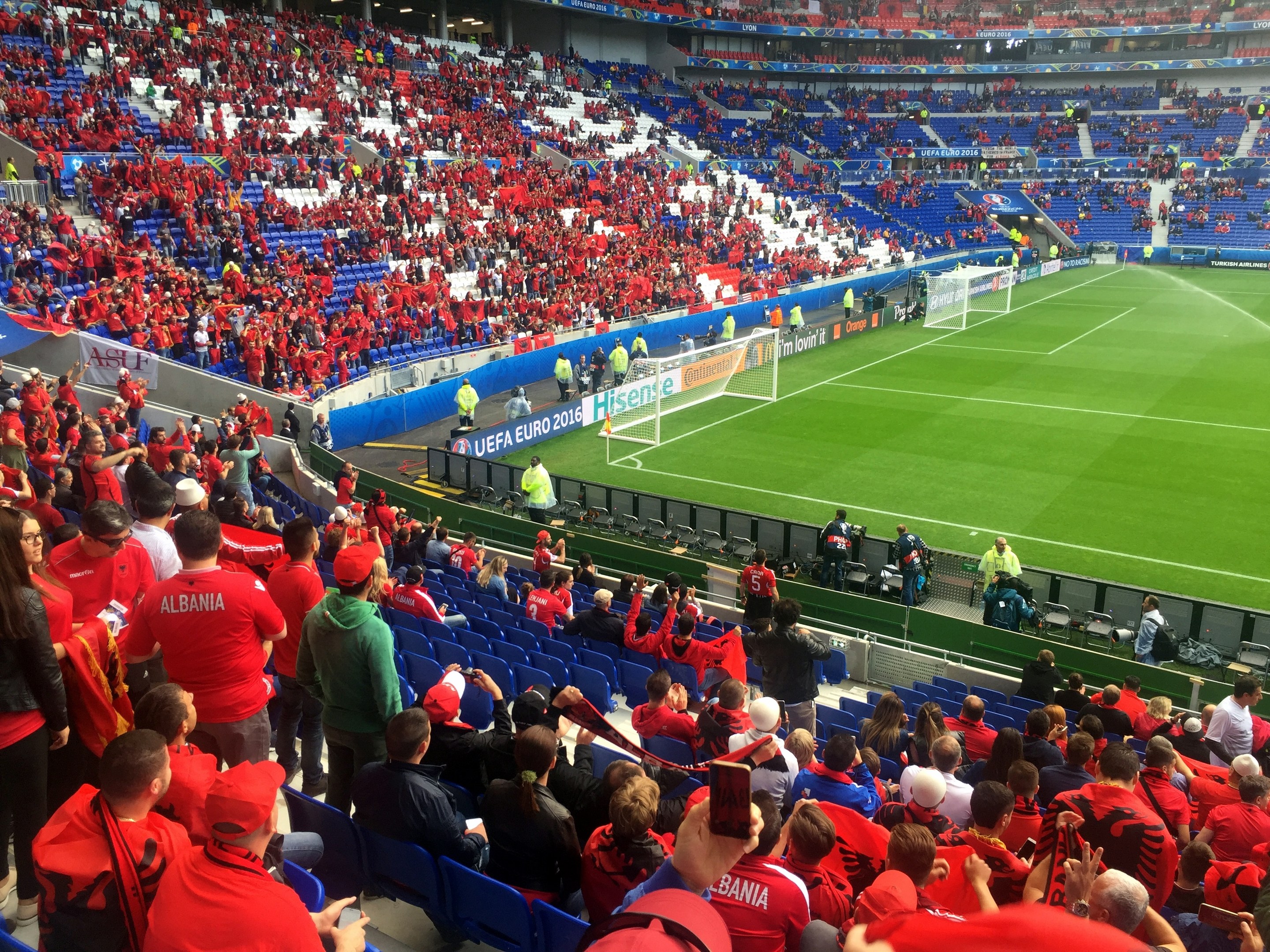 Eine gro├če Menge in einem Stadion schaut einem Fussballspiel zu, mit Menschen, die auf Stühlen sitzen und stehen, Bannern, einem Torpfosten und Deckenlichtern.