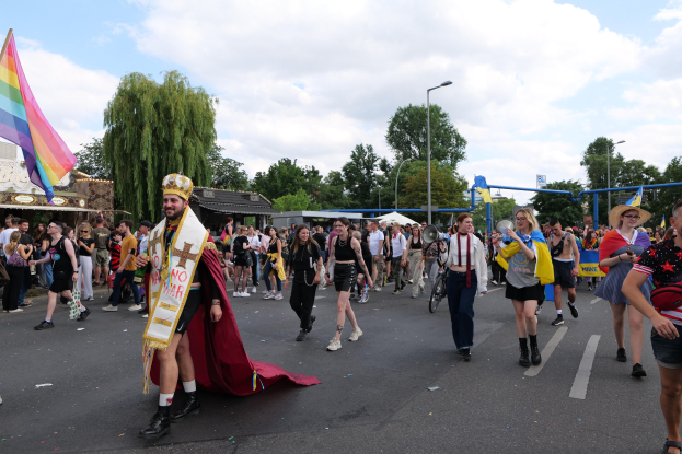 Eine Gruppe von Menschen marschiert bei der Pride Parade 2018, einige mit Musikinstrumenten und andere mit Mützen, die eine Regenbogenflagge tragen, während sie eine Straße mit Laternenpfählen, Bäumen und Schuppen entlanggehen, unter einem bewölkten Himmel.