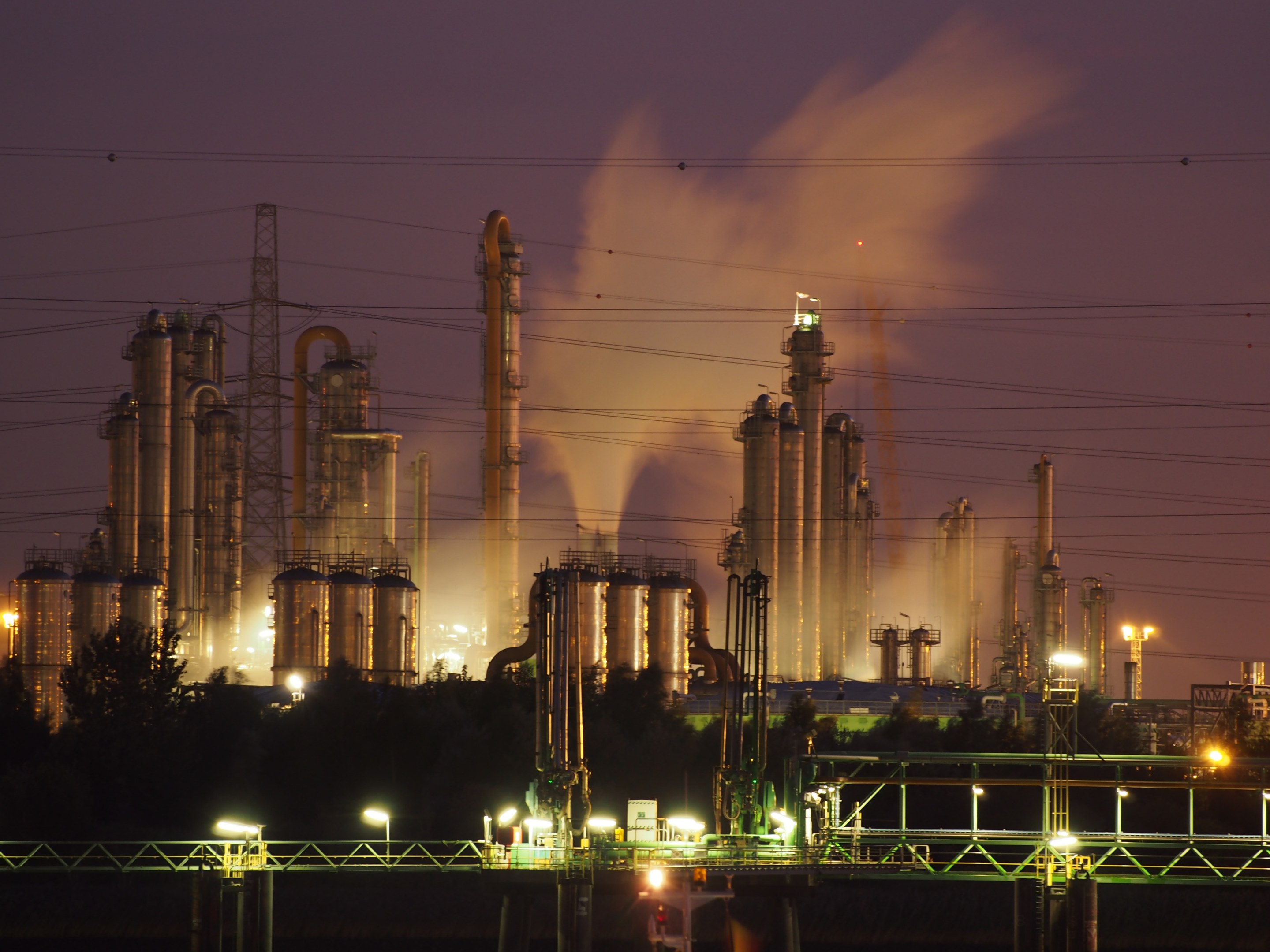 Ölraffinerie bei Nacht mit Rauch, der aus ihr aufsteigt, beleuchtet von umliegenden Lichtern, vor einem sichtbaren Nachthimmel.