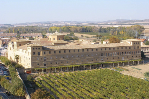 Luftaufnahme eines großen Gebäudes in einer ländlichen Gegend mit Bäumen, Pflanzen und Fahrzeugen auf der Straße, vor einem Hügel- und einem klaren blauen Himmel.