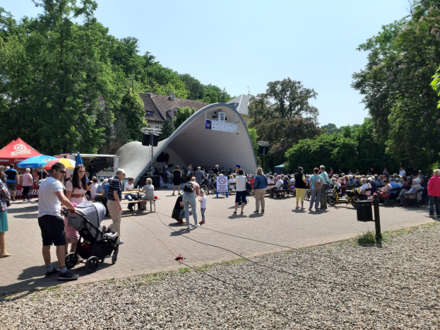 Menschenmenge vor einem Gebäude bei einer Festival-Eröffnung, einige sitzen auf Bänken und andere stehen, mit einem Kinderwagen im Vordergrund, Bäume und Gebäude im Hintergrund, klarer blauer Himmel, Zelte links und ein Mülleimer rechts.