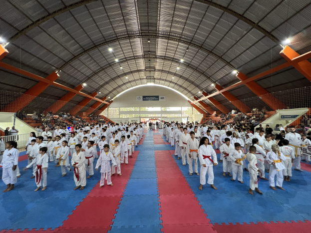 Eine große Gruppe von Menschen auf einer blauen und roten Matte in einer Turnhalle mit anderen auf Treppen sitzend, unter beleuchteter Decke und Schildern im Hintergrund, die auf ein Taekwondo-Event hinweisen.