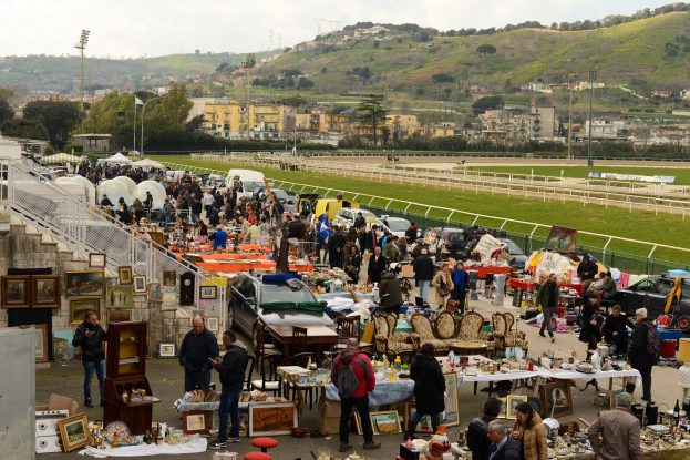 Eine große Gruppe von Menschen auf einem Outdoor-Flohmarkt mit Tischen, auf denen Gegenstände wie Foto Rahmen und Stühle ausgelegt sind, umgeben von geparkten Fahrzeugen, Geländern, Treppen, Bäumen, Gebäuden, Laternenmasten, Hügeln und einem bewölkten Himmel.