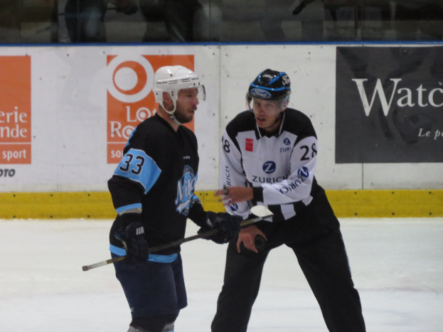 Zwei Männer in Helmen und Eishockey-Ausrüstung spielen Eishockey auf einem Eis, mit einer Wand und Zuschauern im Hintergrund.