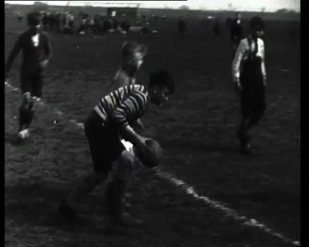Schwarzes und weißes Foto einer Gruppe junger Männer, die Rugby auf einem Feld spielen, einer hält den Ball.