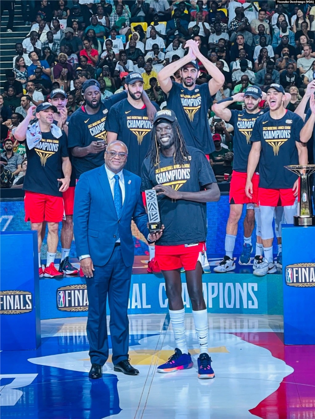 Eine Gruppe von Männern auf einem Basketballfeld feiert mit Trophäen, während andere im Hintergrund sitzen und zwei weitere Trophäen auf Podesten rechts stehen.