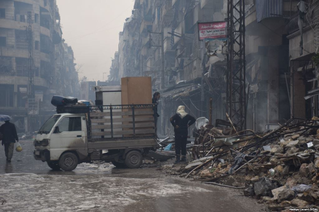 Ein Lastwagen fährt auf einer Straße neben einem Schutthaufen, mit Menschen, die auf der Straße gehen, einer Person mit einem Schirm und Gebäuden, Pfählen und Schildern mit Text im Hintergrund bei klarem Himmel.