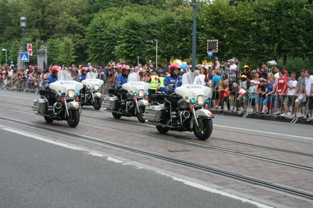 Eine Gruppe von Polizeibeamten auf Motorrädern fährt eine Straße mit Geländern, Laternen, Schildern und Bäumen entlang, während sie von einer Menge Menschen am Straßenrand beobachtet werden.