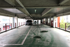 Ein mehrstöckiges Parkhaus mit geparkten Autos, das von Geländern, Säulen, Deckenleuchten, Rohren und umliegenden Bäumen umgeben ist, mit Stadtgebäuden und Fenstern im Hintergrund.