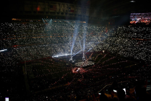 Eine gro├če Menge Menschen in einem Stadion bei Nacht, beleuchtet von hellen Lichtern, mit einem Bildschirm auf der rechten Seite, der wahrscheinlich die Ergebnisse der Super Bowl XLVII-Halbzeitshow anzeigt.