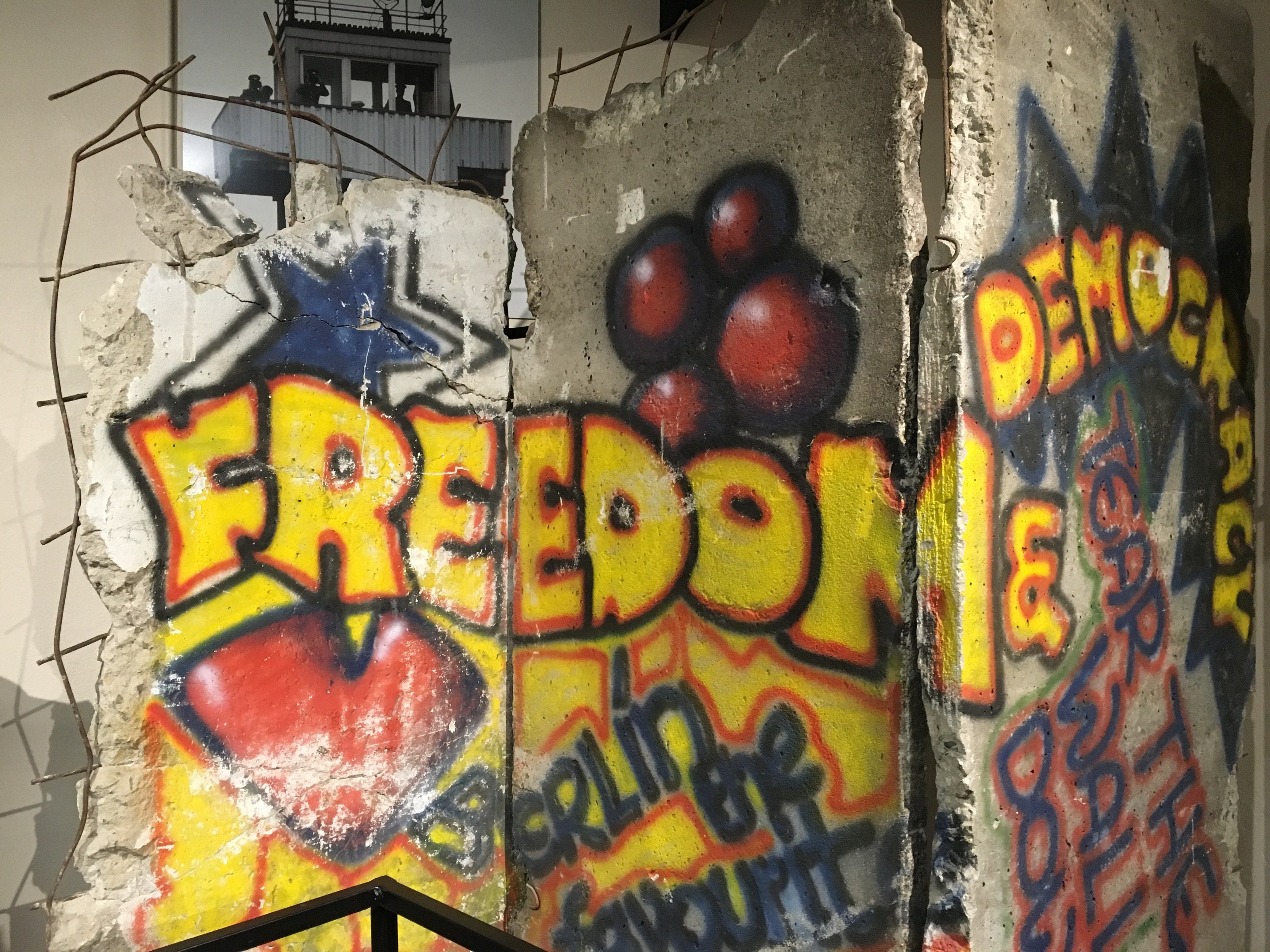 Eine Wand mit Graffiti, das in großen schwarzen Buchstaben 'Freiheit und Demokratie' lautet, ein Geländer unten und ein Gebäude mit Fenstern im Hintergrund.