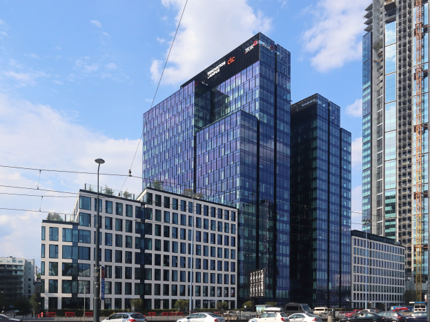 Zentrale der Deutschen Bank in Frankfurt, Deutschland, ein großes mehrstöckiges Gebäude mit zahlreichen Fenstern, umgeben von städtischer Infrastruktur einschließlich Straßenlaternen, Elektroanschlüssen, Elektrokabeln, Kraftfahrzeugen auf der Straße, Büschen, Bäumen und einem wolkenverhangenen Himmel.