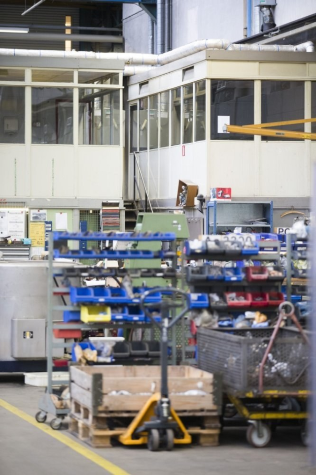 Ein großes Lagerhaus voller verschiedener Geräte, einschließlich Wagen, Schränken und einer Kiste, mit einer Wand aus Glasfenstern und Rohren im Hintergrund.