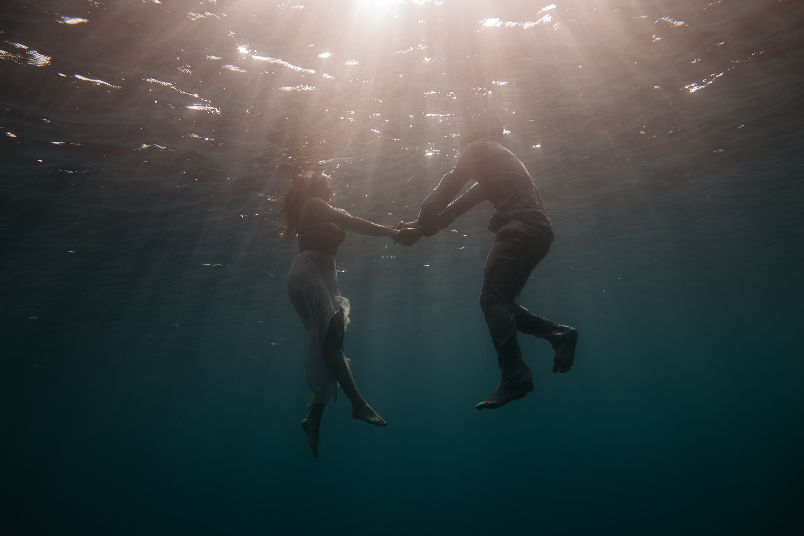 Zwei Personen, die sich unter Wasser in einem Ozean mit einfallenden Sonnenstrahlen an den Händen halten.