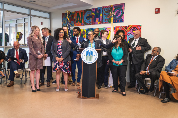 Gruppe von Menschen vor einem Podium mit einem Mikrofon und einer Tafel mit der Aufschrift "Sancho Stadtrat tritt dem Stadtrat bei", mit sitzenden und stehenden Teilnehmern, Fahnen, Feuerlöscher und Deckenleuchten im Hintergrund.