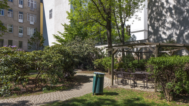 Ein Park mit einem grünen Mülleimer, Gras, Pflanzen, Bäumen, Gebäuden mit Fenstern, verstreuten Stühlen und Tischen sowie einer Hütte im Hintergrund.