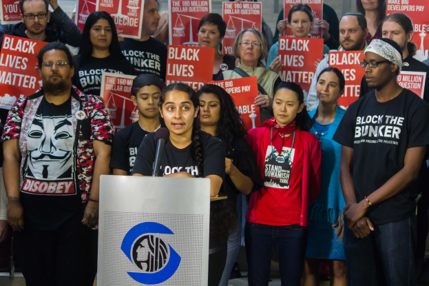 Eine Gruppe von Menschen mit "Black Lives Matter"-Schildern vor einer Menge, mit einer Frau an einem Rednerpult in der Nähe einer Wand.