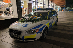 Ein mit Blumen und Blättern geschmückter Polizeiwagen parkt nachts vor einem Geschäft mit Glaswänden, wo Schaufensterpuppen in Kleidern und Informationsschilder zu sehen sind.