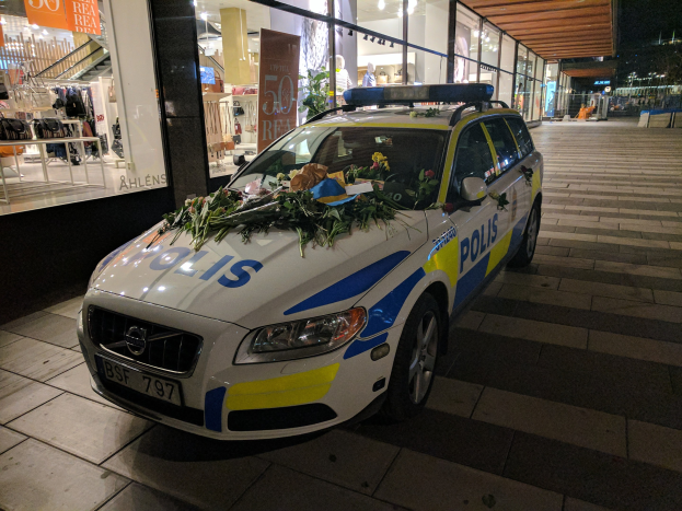 Ein mit Blumen und Blättern geschmückter Polizeiwagen parkt nachts vor einem Geschäft mit Glaswänden, wo Schaufensterpuppen in Kleidern und Informationsschilder zu sehen sind.