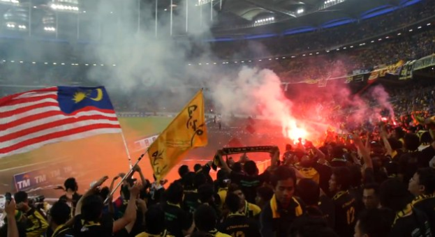 Eine große Menge Menschen in einem Stadion mit brennenden Fackeln in der Luft, einige halten Fahnen und eine Person hält eine Kamera, mit Rauch und Schildern im Hintergrund, die "Malaysia vs Malaysia Live Streaming" anzeigen.