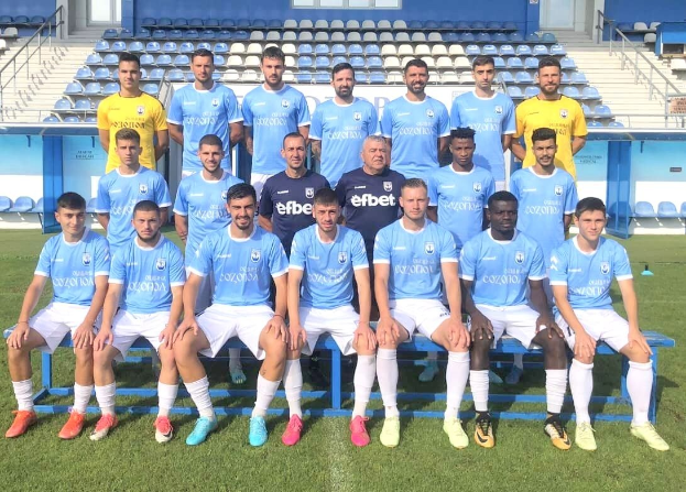 Gruppe von Männern auf einem Fußballfeld, einige sitzen auf Bänken und andere stehen auf dem Rasen, mit Tribünen und Gebäuden im Hintergrund.