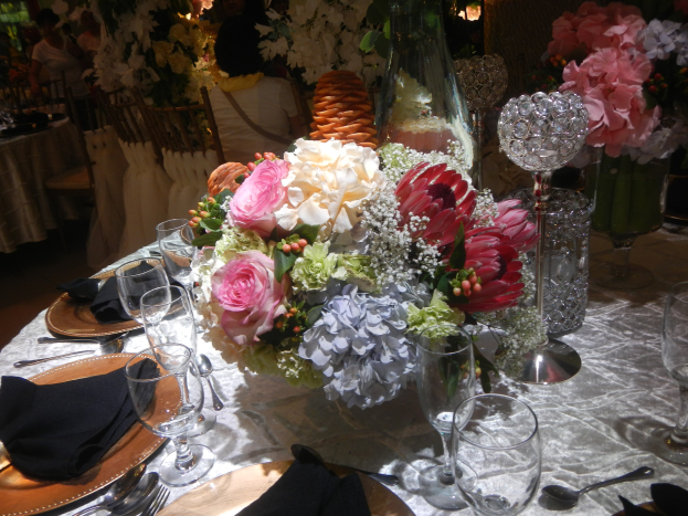Ein formeller Esstisch mit Tellern, Besteck, Gläsern, Servietten und Blumenvasen, mit einem Blumenstrauß in der Mitte und Stühlen und besetzten Tischen im Hintergrund.