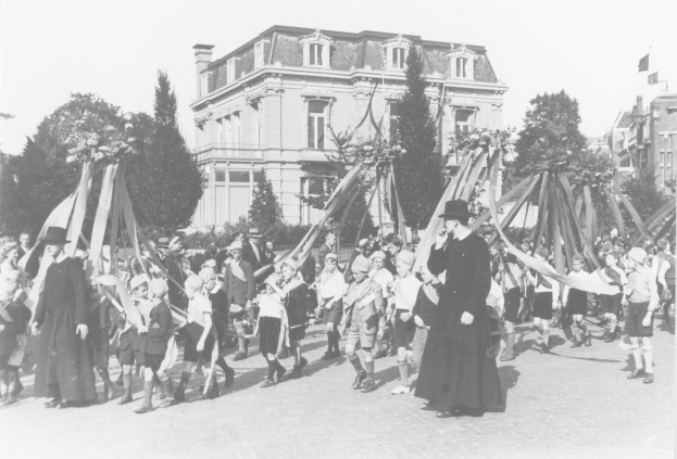 Ein Schwarz-Weiß- Bild einer Gruppe von Menschen, die eine Straße entlanggehen, einige halten Fahnen, mit Hüten, Bäumen, Gebäuden und einem Maibaum im Hintergrund.