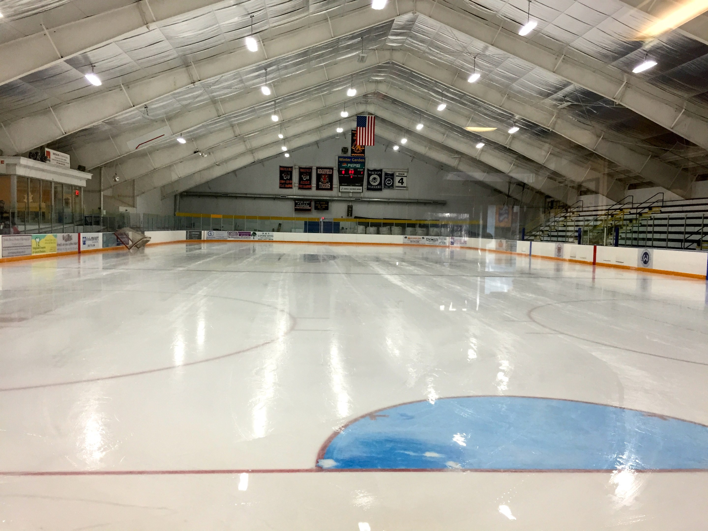 Große Indoor-Eisfläche mit Deckenbeleuchtung, Werbetafeln, Treppen mit Geländern, einer Wand im Hintergrund und einigen Menschen, was auf eine Sportanlage hinweist, die likely für Hockey genutzt wird.
