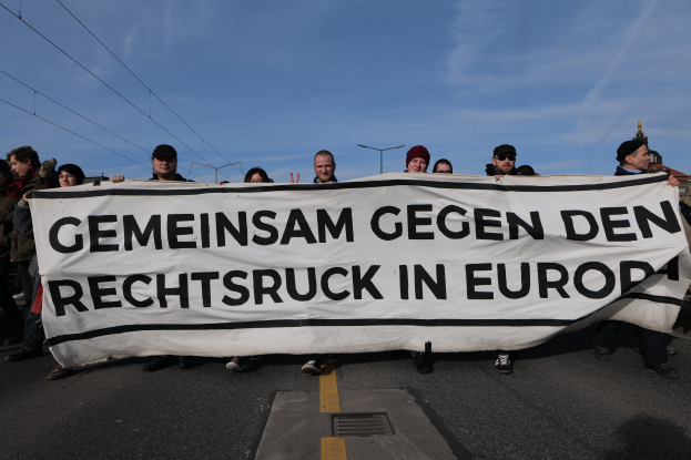 Eine Gruppe von Menschen steht auf der Straße und hält ein Banner mit der Aufschrift "Gemeinsam Gegen den Rechtsruck in Europa" hoch, mit Straßenlaternen, Strommasten, Kabeln, Gebäuden und einem bewölkten Himmel im Hintergrund.