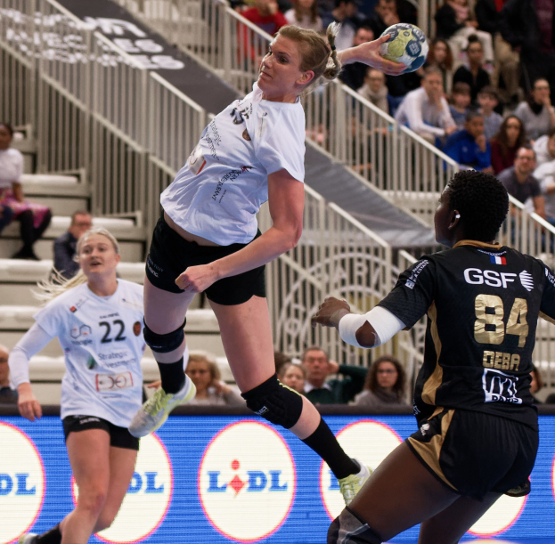 Eine Gruppe von Frauen, die Handball auf einem Platz spielt, mit einem Ball in der Luft, Zuschauern auf Treppen und Geländern und einem "Handball-Weltmeisterschaft 2019 - Tag 2"-Schild im Hintergrund.