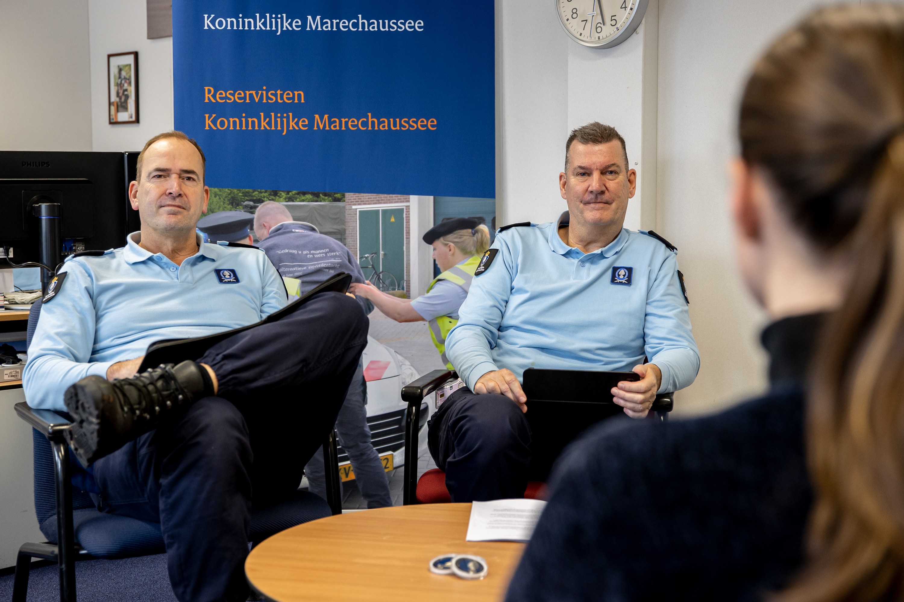 Zwei Polizeibeamte sitzen an einem Tisch mit Papieren, sprechen mit einer Frau; eine Wandbanner, Uhr und Foto Rahmen im Hintergrund, mit einem weiteren Tisch mit Monitor, Tastatur und Maus links.