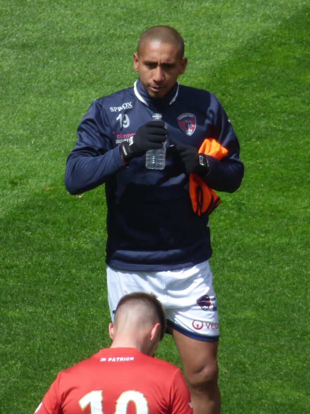 Ein Profisportler in einem Sportdress steht auf einem saftig grünen Feld, in einer Hand hält er eine Flasche und in der anderen ein oranges Tuch.