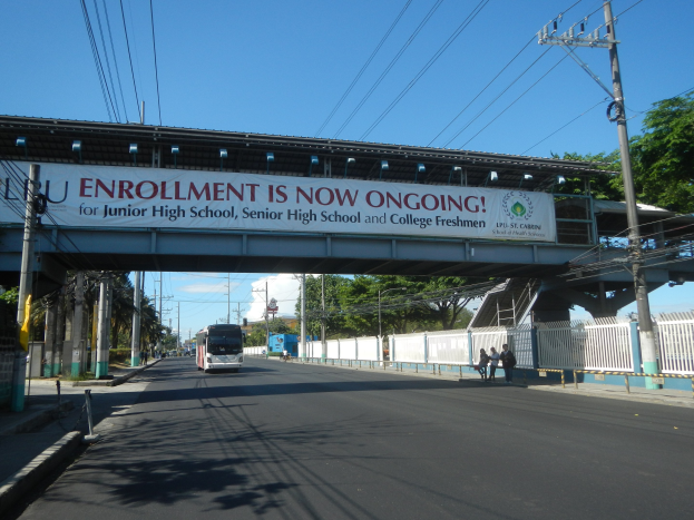 Ein Bus, der unter einer Brücke mit einem "Jetzt Anmelden"-Schild fährt, Menschen, die auf dem Gehweg darunter gehen, Strommasten mit Drähten, Bäume und ein klarer blauer Himmel im Hintergrund.