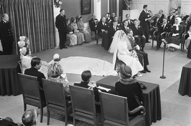 Schwarzes und weißes Foto einer Hochzeitszeremonie mit Menschen, die sitzen und stehen, einem Mann mit Mikrofon, einer Frau mit Blumenstrauß, einem Podium mit Mikrofon, Blumenbouquets, einem Vorhang, einem Foto im Rahmen an der Wand und einer Tür im Hintergrund.
